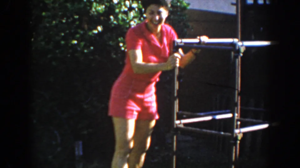 A smiling woman in a pink short suit stands beside a metal exercise frame in a sunlit North Hollywood yard.