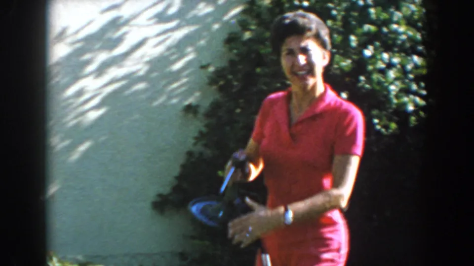 A woman in a pink short suit smiles toward the camera in a North Hollywood yard, holding a dark object near the shrubs and wall.
