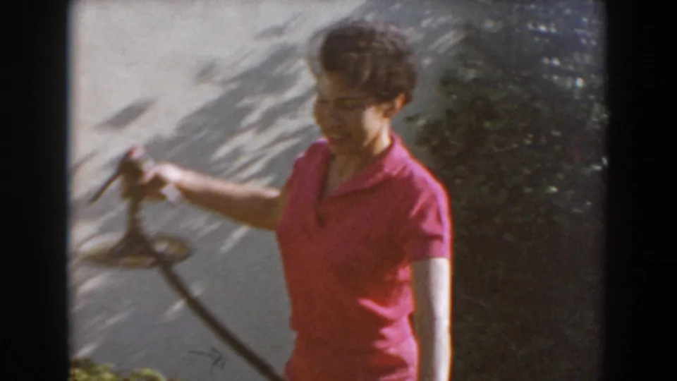 Adult woman in a pink polo holds a garden hose near shrubs in 1962 North Hollywood home-movie footage.