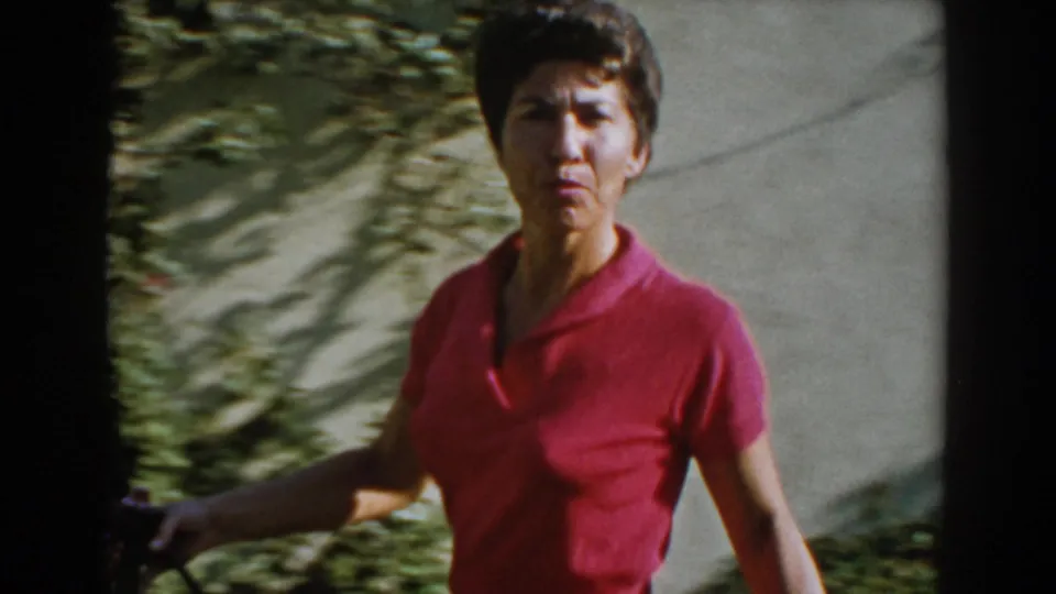 In a North Hollywood garden, an adult woman in a pink shirt looks toward the camera while holding a hose.