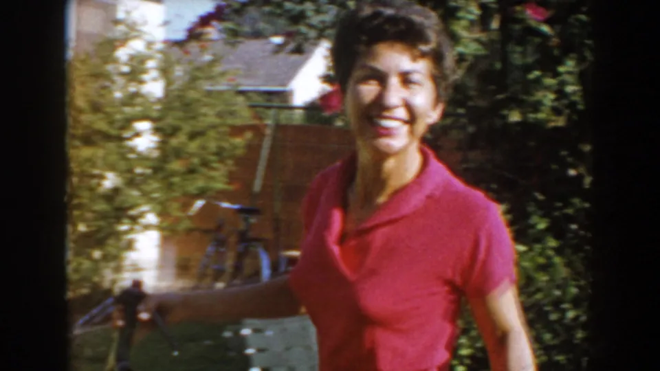 Smiling adult woman in a pink shirt holds a garden hose in a leafy North Hollywood yard, 1962.