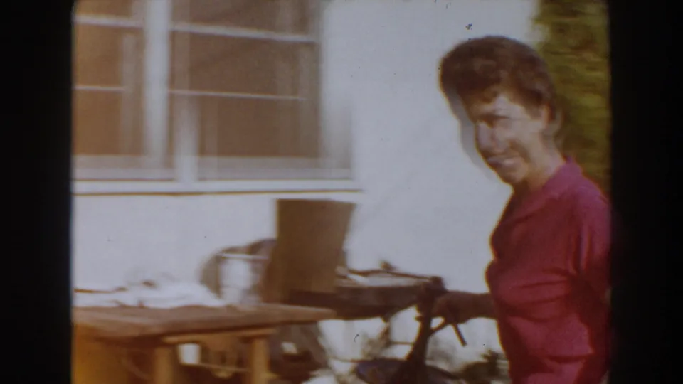 Adult woman in a pink shirt stands with a garden hose near a house exterior and window in archival home-movie footage.