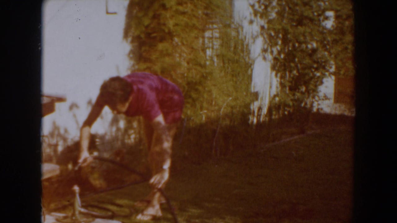 In 1962 North Hollywood home-movie footage, a woman in a purple top bends over a hose or tool in a bright yard.