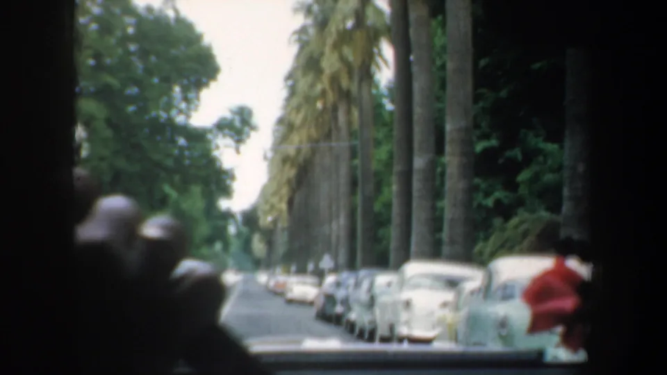 1957 California 8mm frame from inside a moving car, showing a palm-lined street, parked cars, and indistinct passengers or hands near the window.