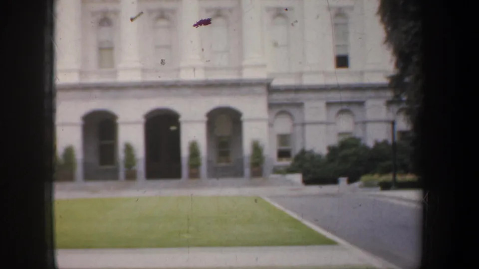 1957 San Francisco frame showing a white building facade with arched entrances, manicured lawn, walkway, shrubs, and roadside motion blur.
