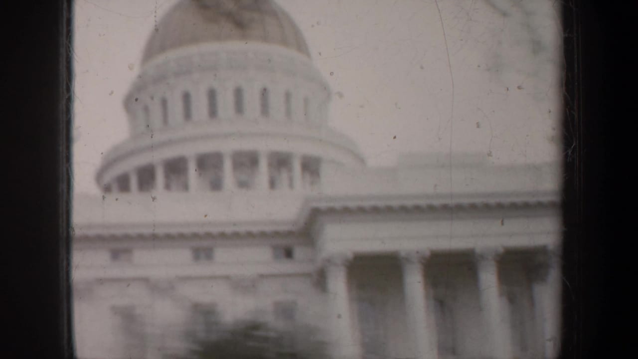1957 8mm frame showing a domed California capitol-style building with columns, trees, and dark vertical framing in the foreground.