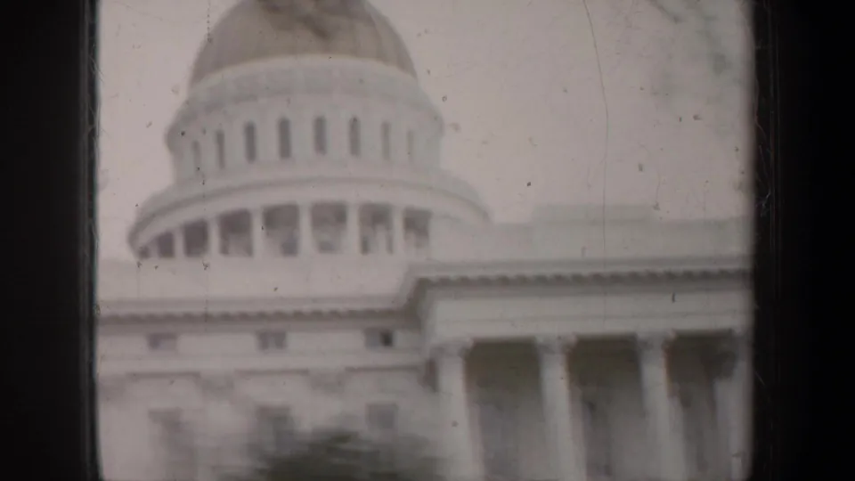 1957 8mm frame showing a domed California capitol-style building with columns, trees, and dark vertical framing in the foreground.