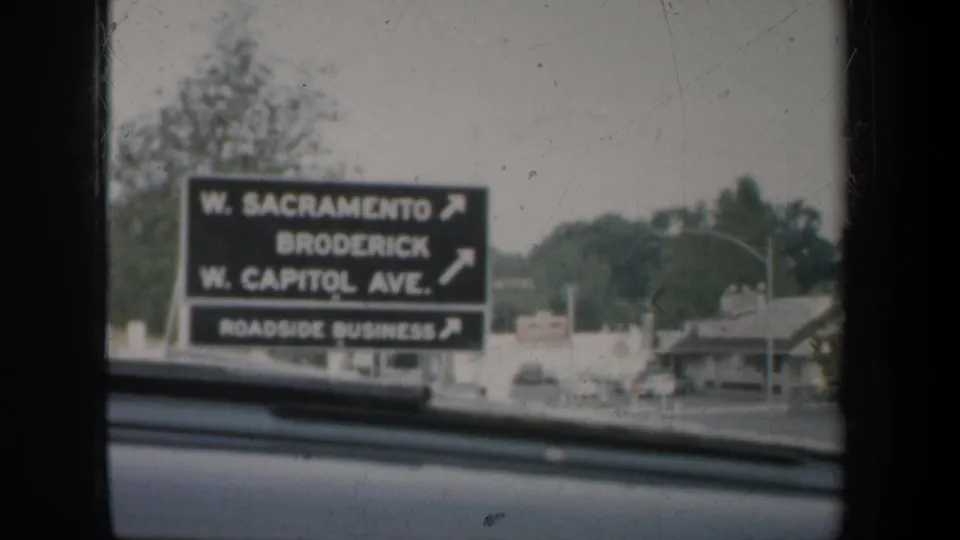 1957 California car-window view of a roadside directional sign listing W. Sacramento, Broderick, W. Capitol Ave., and roadside businesses.