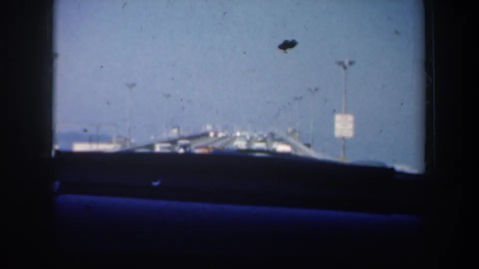 Driver-perspective 1957 San Francisco bridge view with dashboard shadow, roadway lights, distant traffic and a small sign.