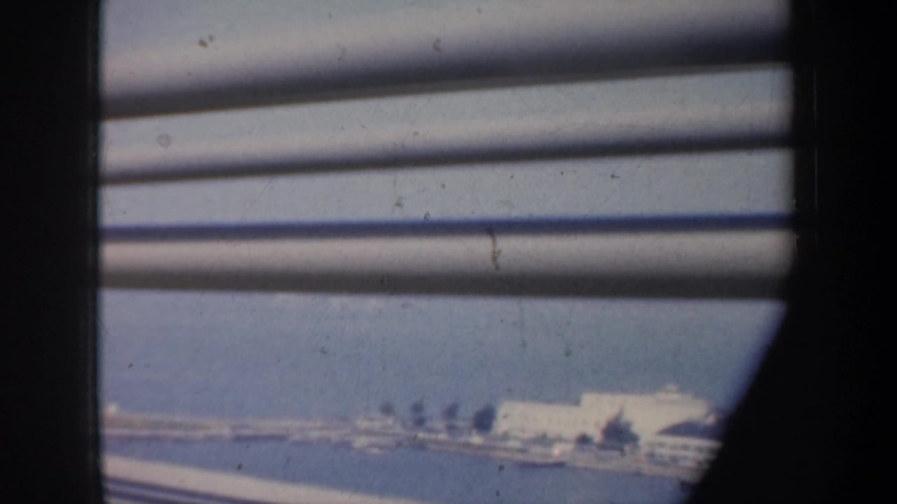 1957 San Francisco coastal view through slats, with blue water, pale shoreline buildings, and soft 8mm film texture.