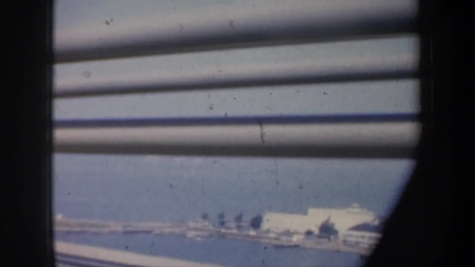 1957 San Francisco coastal view through slats, with blue water, pale shoreline buildings, and soft 8mm film texture.