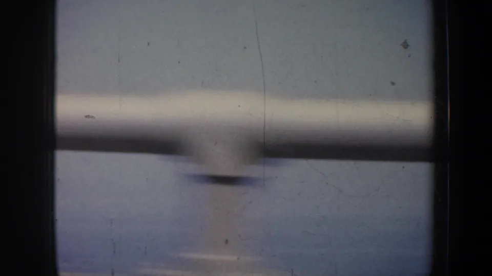 Motion-blurred 1957 travel frame showing window structure, railings, water, and a faint shoreline beyond.