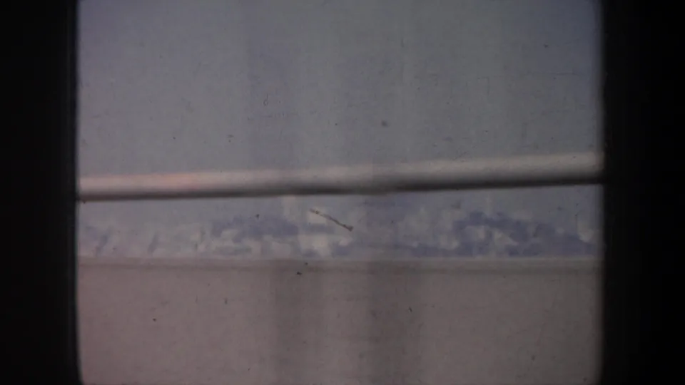 1957 San Francisco 8mm frame showing a muted bridge-side view through a car window, with railing, haze, and distant indistinct scenery.
