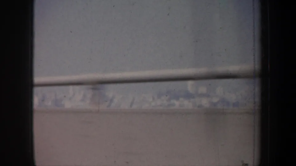 Late-frame 1957 San Francisco bridge-crossing view with a dark window divider, horizontal railing, pale haze, and archival 8mm wear.