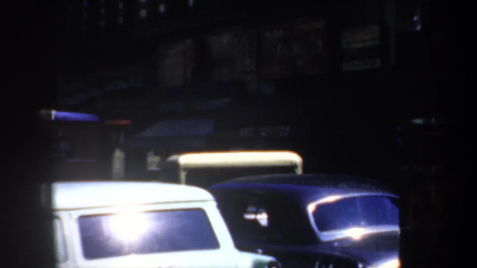 1957 San Francisco street frame with a white station wagon, dark parked cars, glare, and indistinct storefront signage.