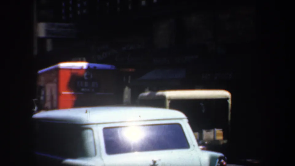 1957 San Francisco 8mm frame showing a white station wagon in the foreground with dark cars and shopfronts behind it.