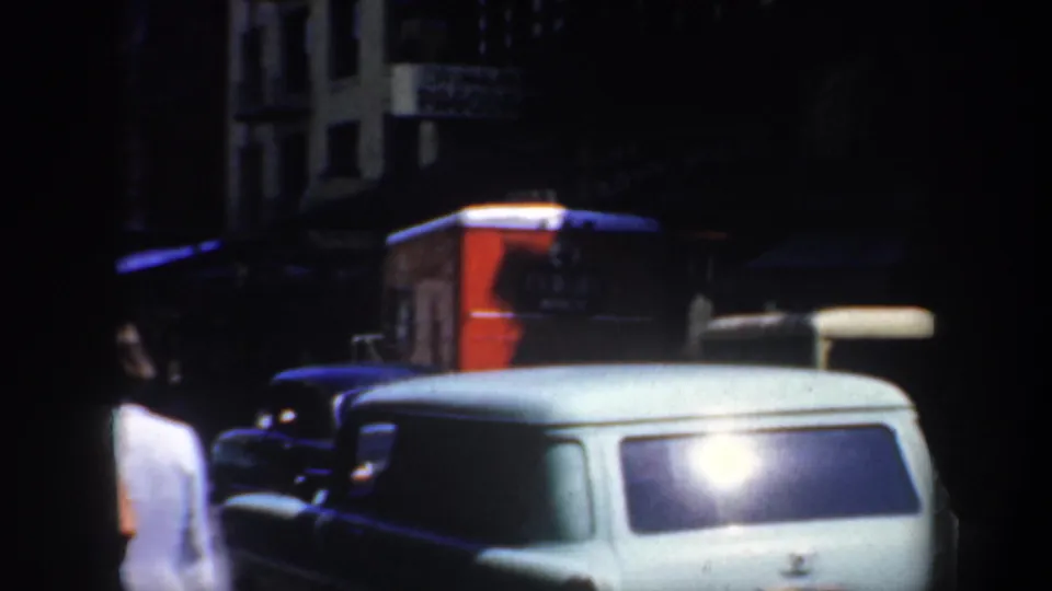 1957 San Francisco frame with a white station wagon in front, dark parked cars, a red vehicle or sign panel, and low-light storefronts.