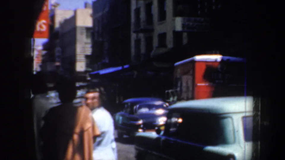 1957 San Francisco Chinatown street view with pedestrians, mid-century cars, dark storefronts, and bright sun beyond the sidewalk.