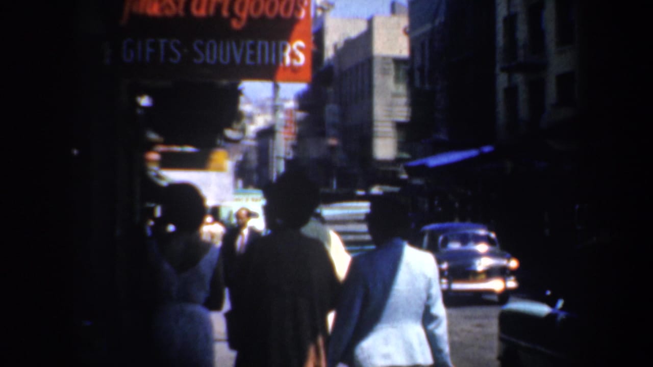 People walk beneath a red gifts and souvenirs sign on a city sidewalk.