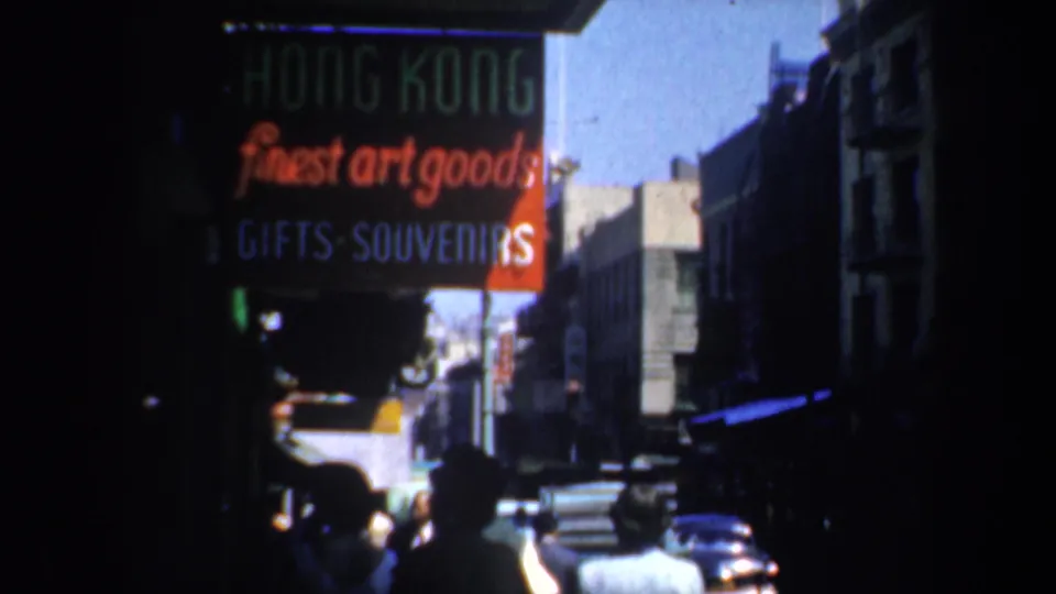 1957 Chinatown street view showing a red Hong Kong art-goods and souvenirs sign, pedestrians, storefronts, and mid-century cars.