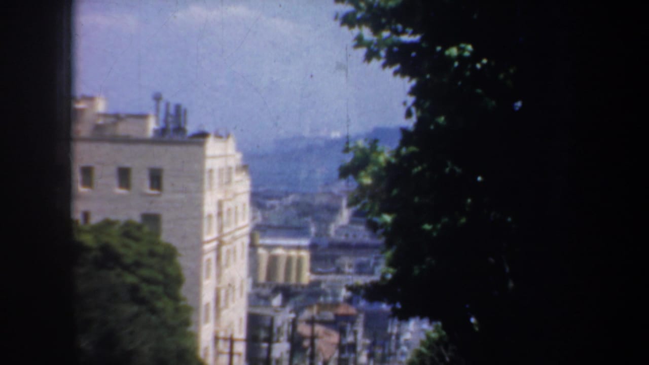 Steep San Francisco street seen between dark window edges, with buildings, trees, and distant hills.