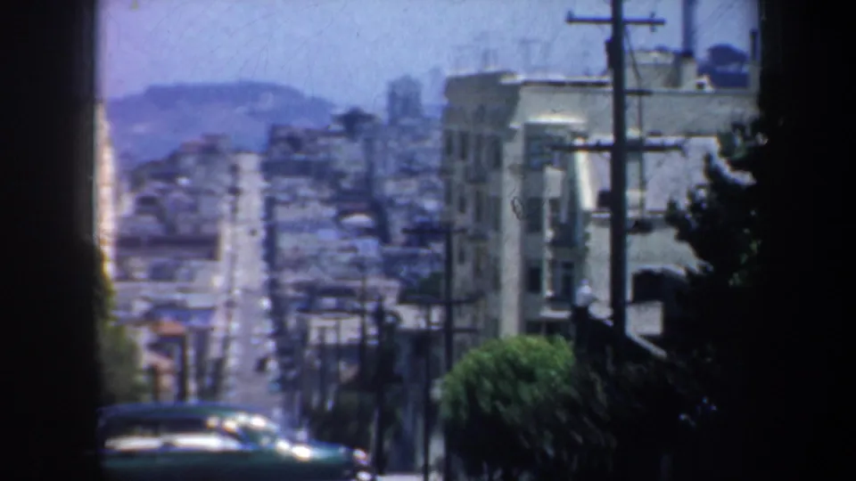 1957 San Francisco street view looking downhill past utility poles, apartment buildings, trees, and mid-century cars.