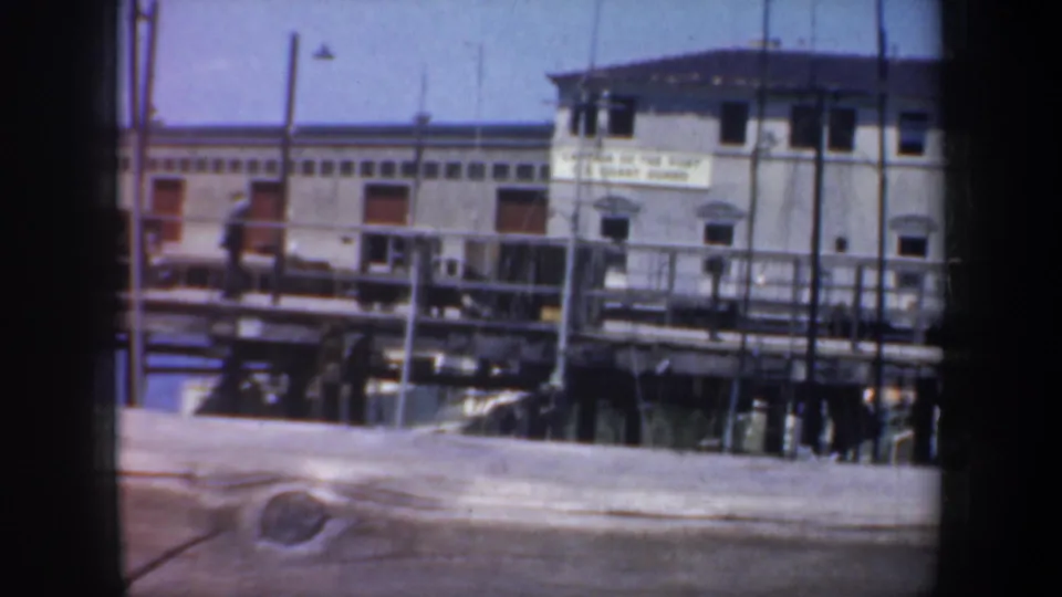 1957 San Francisco waterfront view with a low building, pier railings, a partially readable sign, and soft 8mm color.