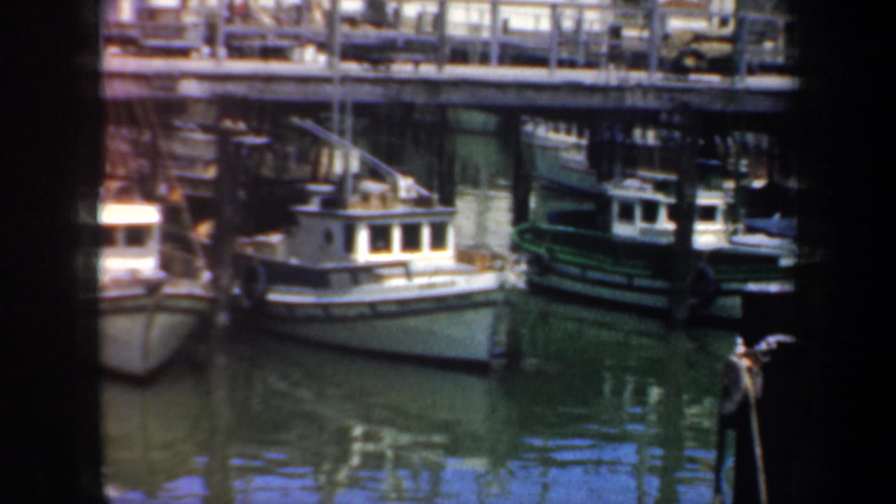 1957 San Francisco harbor view with small moored boats, dock railings, pilings and reflections on calm water.