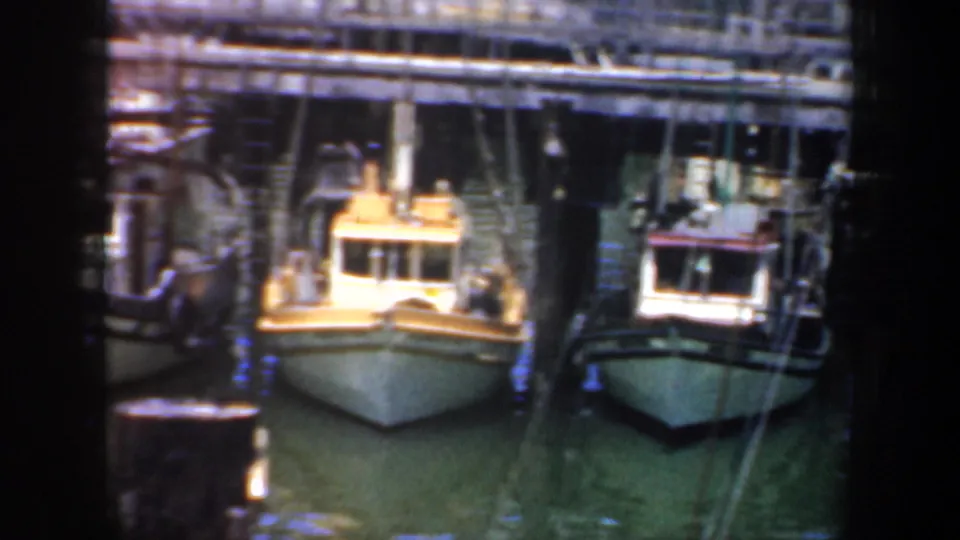 1957 harbor footage shows the bows of small moored boats below a wooden dock, with soft focus and aged 8mm color.