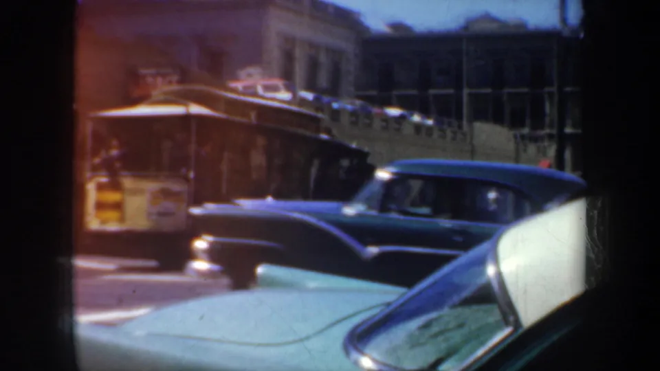Downtown San Francisco traffic scene with a dark classic car, trolley, foreground auto hood, and multi-story buildings in the background.