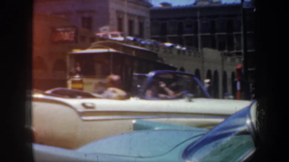 Mid-century convertible traffic in 1957 San Francisco, with a trolley, arched streetscape, and distant pedestrians partly visible.