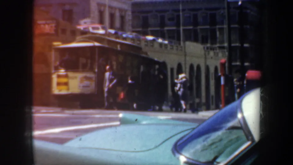 1957 San Francisco downtown street with pedestrians near a trolley, foreground cars, arched architecture, and sunny rush-hour traffic.