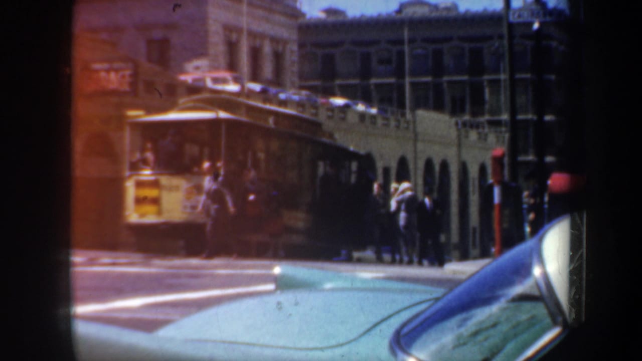 1957 San Francisco street view with a yellow trolley, pedestrians, mid-century buildings, and a light blue car in the foreground.