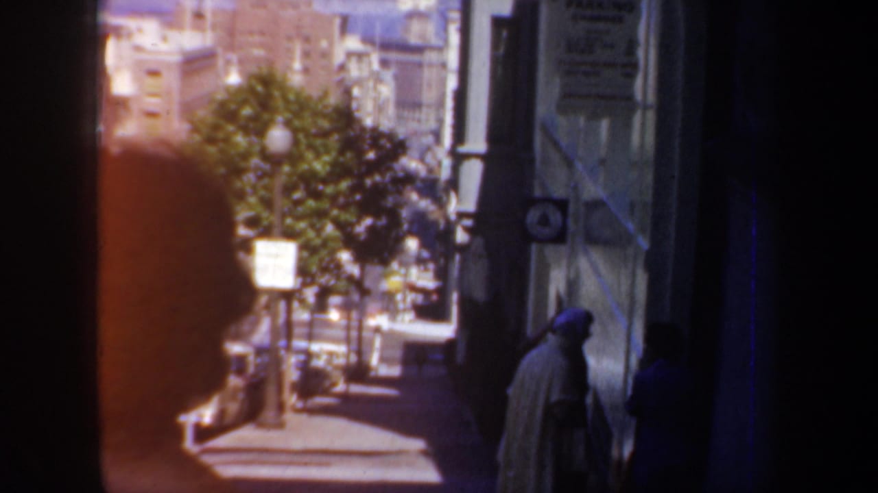 Sidewalk view with pedestrians, a tree, parked cars, and building fronts.