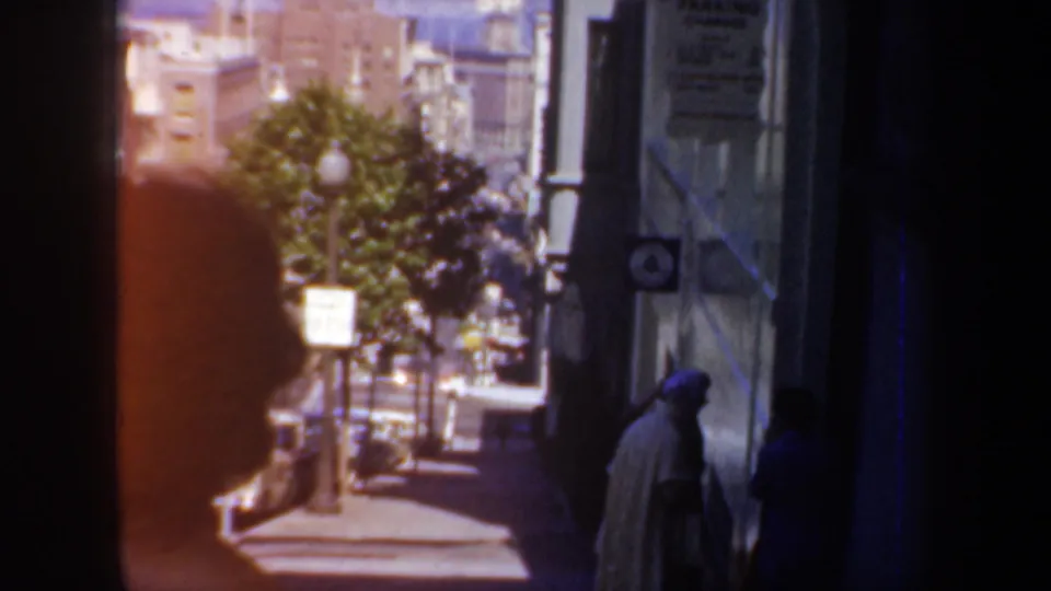 1957 San Francisco sidewalk scene with pedestrians, a street tree, parked cars, and older building facades in soft 8mm color.