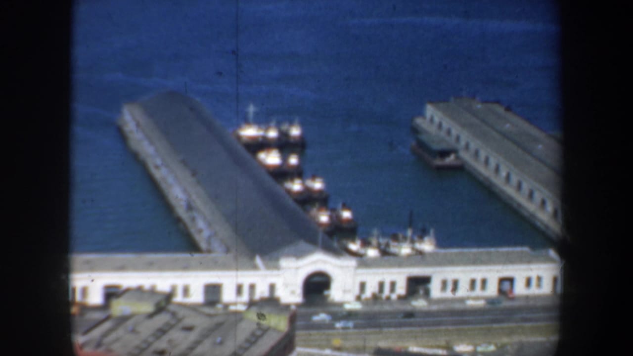 High-angle view of white waterfront buildings, long piers, docked boats, and blue bay water.