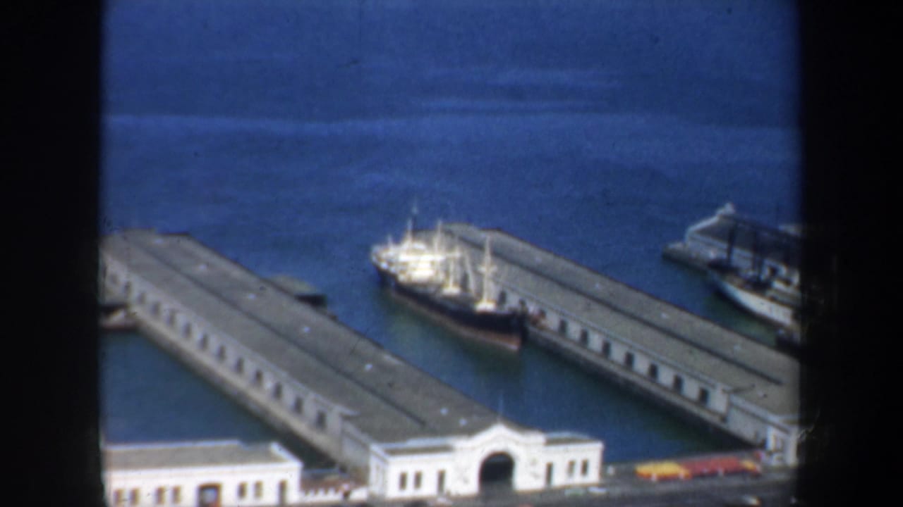 Long pier buildings extend into blue water with a white cargo ship docked between them.