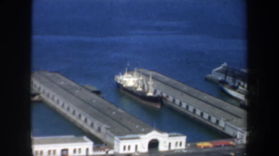 1957 San Francisco port view with long pier sheds, a white and dark-hulled ship docked at the waterfront, and bay water beyond.