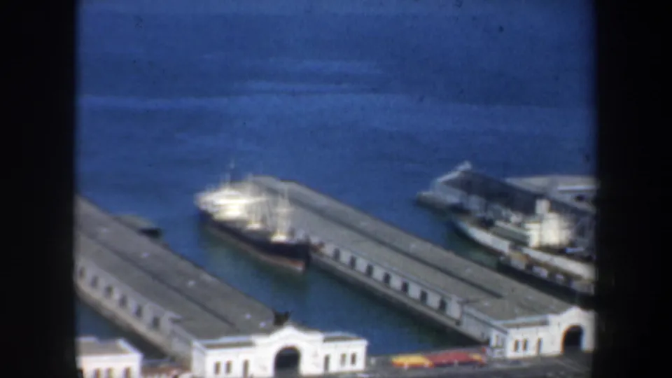 1957 San Francisco harbor frame showing pier sheds, a docked ship, blue water, and small vehicles along the waterfront edge.