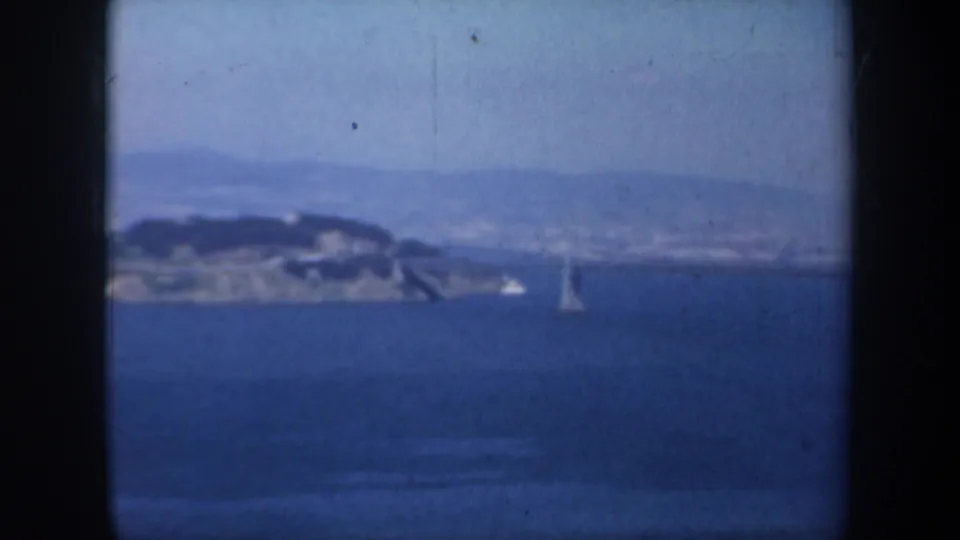 1957 San Francisco Bay view with a distant hilly island, hazy shoreline, blue water, and a small sailboat in the foreground.