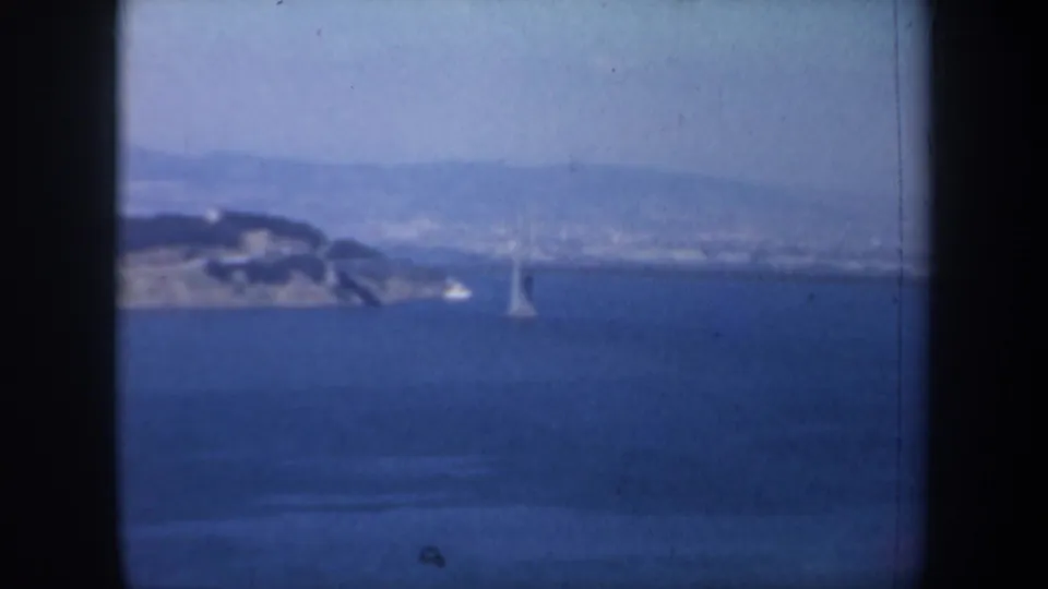 1957 San Francisco Bay overlook with blue water, a sailboat, distant shoreline, and a bridge tower seen through haze.