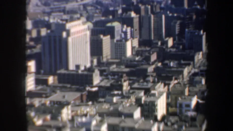 San Francisco skyline from a high vantage point, with tall mid-century buildings, rooflines, and a hazy distance visible through dark framing.