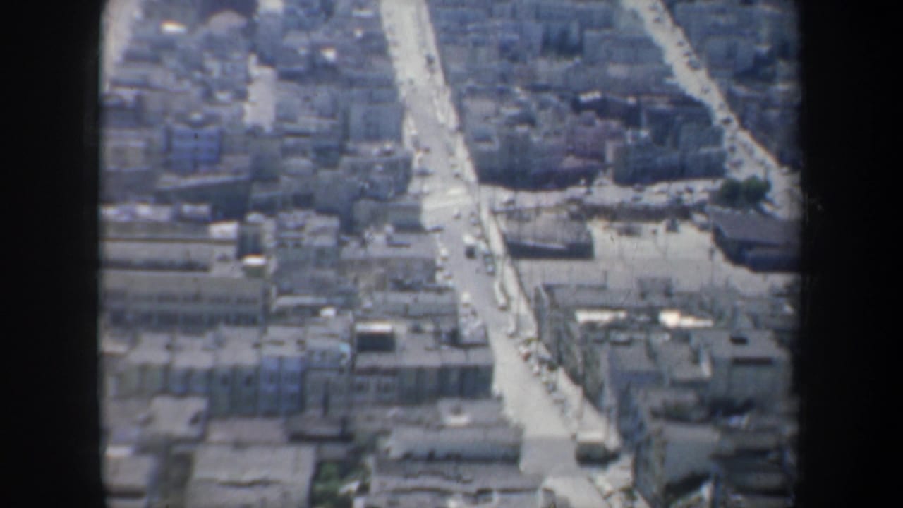 Aerial view over dense city blocks with a bright straight street and small cars.