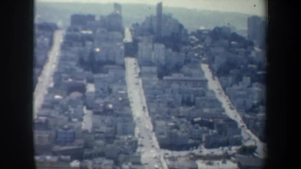 1957 San Francisco aerial frame with long streets, dense buildings, and a hazy horizon in soft home-movie focus.