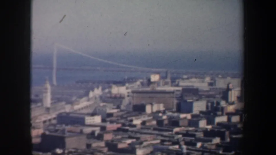 A 1957 home-movie frame shows the San Francisco waterfront, piers, and Bay Bridge beyond dense city blocks.