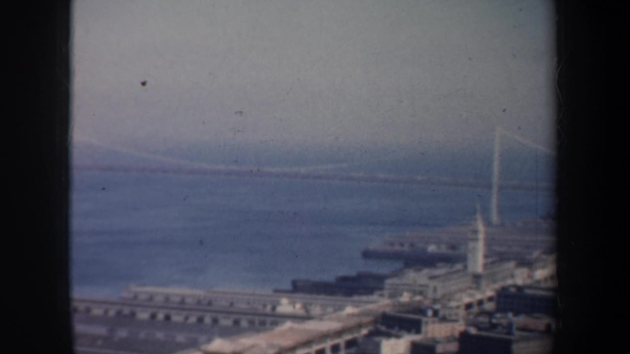 1957 8mm home-movie frame looking over San Francisco Bay toward a suspension bridge, waterfront piers, and a small sailboat.