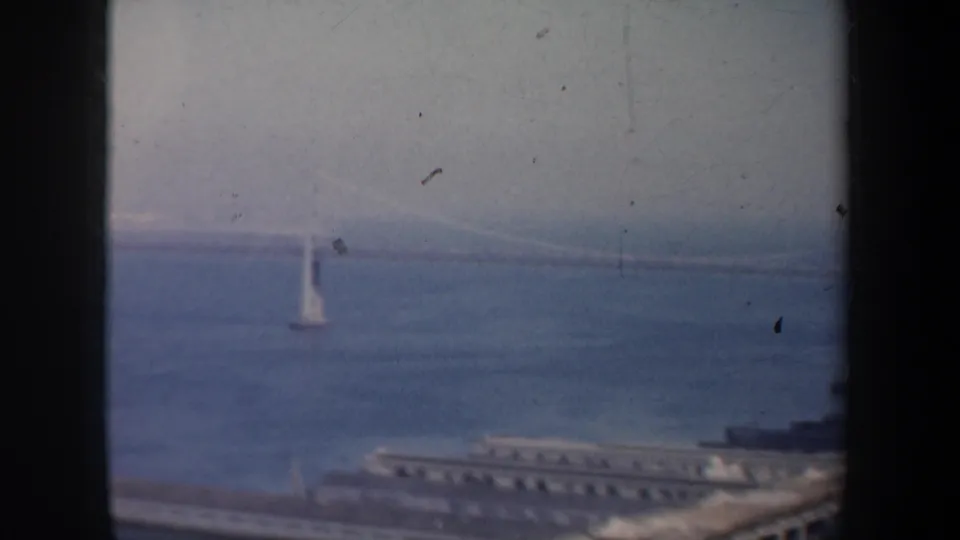 1957 San Francisco Bay frame with a distant suspension bridge, a white sailboat on the water, and waterfront structures below.