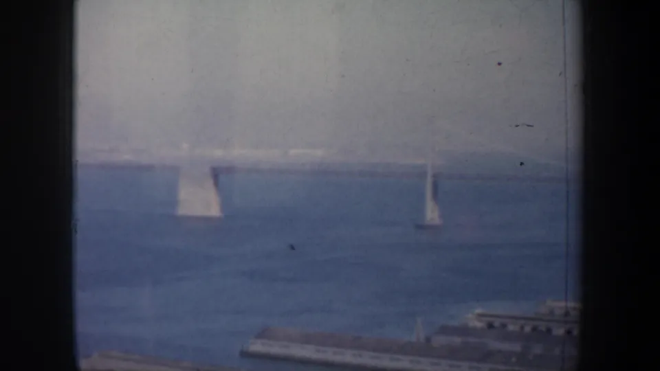 Soft 8mm frame from San Francisco Bay showing bridge towers across open water with piers and shoreline structures at the lower edge.