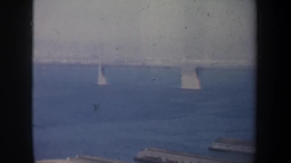 1957 home-movie frame over San Francisco Bay, with small sailboats on the water, faint bridge elements, and piers along the lower edge.