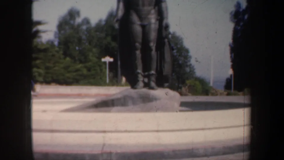 1957 San Francisco frame showing the lower portion of a bronze standing statue, stone pedestal, plaza paving, and nearby trees.
